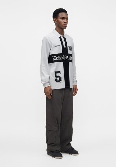 Jeune homme debout, portant un polo blanc et noir à manches longues avec un texte et le numéro 5, un pantalon cargo foncé et des baskets noires.