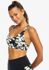 Sujetador deportivo con un patrón floral en negro, blanco y azul, con tirantes anchos y escote redondeado. Tejido texturizado y diseño ajustado.