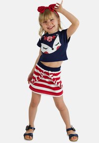 Granatowy t-shirt z grafiką lisa i kwiatowymi detalami; spódnica w czerwono-białe paski z falbaną na dole. Niebieskie sandały dopełniają całość.