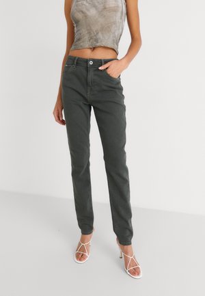Person iført mørkegrå bukser med lige ben, grå ærmeløs cropped top og hvide højhælede sandaler med stropper, stående mod en ensfarvet baggrund.
