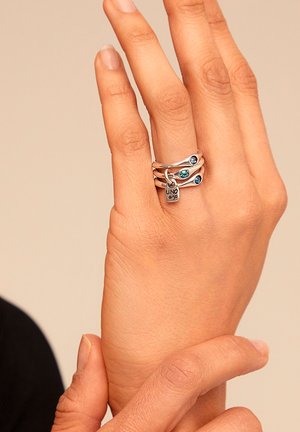Tres anillos plateados apilados en una mano, con diversas piedras azules y un pequeño colgante en forma de candado. Superficie lisa con acabado pulido.