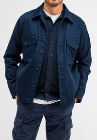 Navyblaue Baumwoll-Arbeitsjacke mit zwei Brusttaschen, Knopfverschluss und passendem dunklen Hemd darunter. Hat ein schlichtes weißes Hemd am Grund.