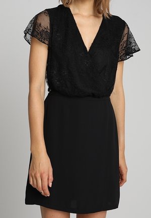 Kobieta w czarnej sukience z krótkimi koronkowymi rękawami w kwiatowy wzór i dekoltem w serek, przód, długość powyżej kolana.