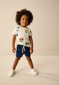 Barn som bär en krämfärgad t-shirt med färgglada biltryck, ihop med mörkblå shorts med dragsko. Vita sneakers fulländar outfiten.