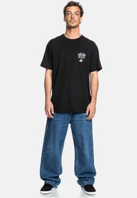 Black cotton t-shirt featuring white graphic with checkered flags, paired with loose blue denim jeans and black sneakers with white soles.