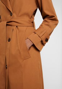 Abrigo trench marrón con cintura ceñida, botones decorativos y bolsillos laterales, confeccionado en un tejido liso y texturizado con diseño de mangas entalladas.