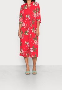 Robe fleurie rouge avec une patte de boutonnage à l'avant, une taille nouée, des manches trois-quarts et des motifs de feuilles vertes. Associée à des sandales à talons vertes.