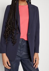 Kvinna med långt flätat hår iklädd en marinblå blazer, korallfärgad t-shirt och mörkblå högmidjade byxor, som står mot en enfärgad bakgrund.