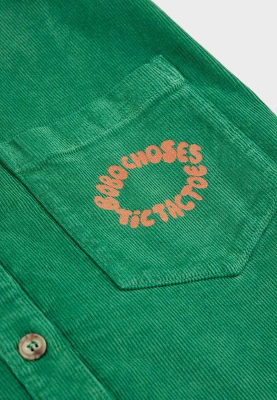 Grüne Cordhemd mit einer Brusttasche, auf der orangefarbener, kreisförmiger Text mit "Bobo Choses Tictactoe" zu sehen ist. Sichtbares Knopfdetail.