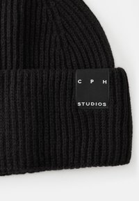 Melnā ribota adīta cepure ar saliektu aproci, uz kuras ir taisnstūra melns uzlīmes gabals ar uzrakstu "CPH STUDIOS" baltajās burtiņās.