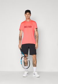 Lys korall t-skjorte med 'BORG' logo, kombinert med svarte shorts. Hvite joggesko og en svart caps fullfører det sporty utseendet. Holder en racket.