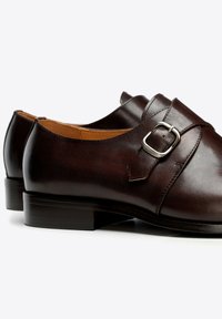 Dunkelbraune Ledermonks mit silberner Schnalle und niedrigem Absatz, von der Seite auf weißem Hintergrund gezeigt.