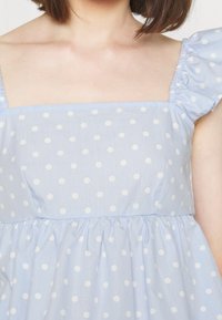 Woman wearing a light blue dress with white polka dots and ruffled short sleeves, cropped to show upper torso and neckline.