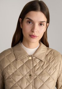 Beige gewatteerde jas met een ruitpatroon, gouden knopen en een spitse kraag, gedragen over een lichtgeribbelde trui. Glad, glanzend textuur.