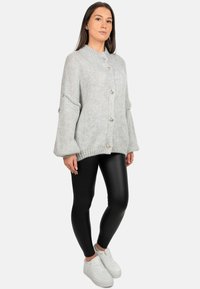 Cardigan lavorato a maglia grigio chiaro con collo alto, maniche oversize e bottoni argentati, abbinato a leggings neri e sneakers bianche.