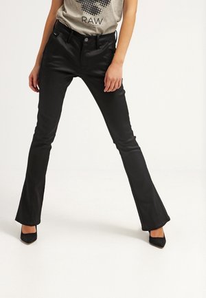 Kobieta ubrana w obcisłe, czarne spodnie z rozszerzanymi nogawkami i czarne buty na obcasie z ostrym noskiem, stojąca z nogami rozstawionymi na białym tle.