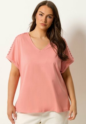 Femme aux longs cheveux bruns portant un haut rose ample à col en V avec des manches courtes et des détails en dentelle le long des épaules.