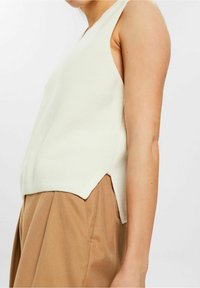 Femme portant un haut en tricot crème sans manches avec des fentes sur les côtés associé à un pantalon marron taille haute sur fond blanc.