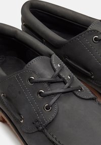 Zapatos náuticos de cuero negro con cordones grises, ojales de metal, detalles cosidos y cuellos acolchados sobre suelas de goma marrón.