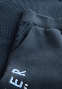 Mørkeblå sportbukser med glat stof og ribbet tekstur på linningen og lommerne; med hvid broderet logodetalje.