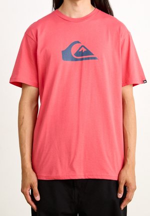 Rotes Kurzarm-T-Shirt mit einem dunkelblauen Wellen- und Berglogo auf der Brust, getragen von einer Person mit langen dunklen Haaren und schwarzer Hose.