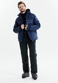 Marineblå jakke med hætte, der har lyse grå striber, lynlåslommer og en glat tekstur. Sammen med sorte bukser og skistøvler.