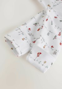 Un tissu blanc présente des illustrations festives, incluant des ornaments de Noël, des cadeaux et des décorations, sur les poignets des vêtements.