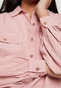 Camicia button-up rosa chiaro con maniche lunghe e voluminose, tasche sul petto e bottoni traslucidi. Tessuto morbido con una texture leggermente stropicciata.