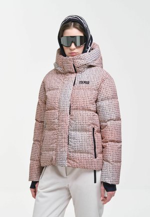 Personne portant une doudoune à capuche rose et blanche à motifs, des lunettes de ski noires et un pantalon blanc sur un fond uni.