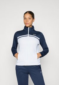 Jaqueta de fecho branco e azul marinho com gola alta, apresentando uma larga faixa horizontal branca e o logótipo no lado esquerdo do peito. Material suave, corte relaxado.