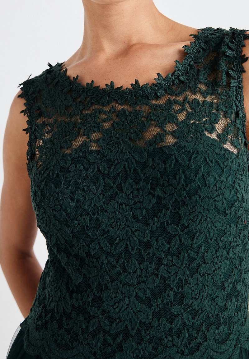 Mörkgrön spetsblus med en texturerad blommönstring, scalloped halsringning och genomskinliga områden som framhäver den intrikata detaljeringen.