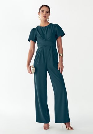 Mujer de pie que lleva un mono azul verdoso oscuro con mangas abullonadas, sosteniendo un bolso de mano dorado, pulsera dorada de brazalete, pendientes y sandalias de tacón alto.