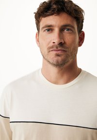 Světlý béžový tričko s kulatým výstřihem a černým vodorovným pruhem přes hruď, který má hladkou, jemnou texturu.