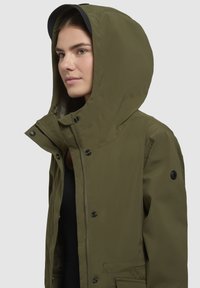 Olive zöld kapucnis dzseki cipzárral és patentos záródással, fekete béléssel és egy folttal a bal ujján.