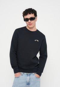 Fekete kereknyakú pulóver bordázott mandzsettával és szegéllyel, fehér hímzett logóval. A modell világoskék, kopott farmernadrágot és napszemüveget visel.