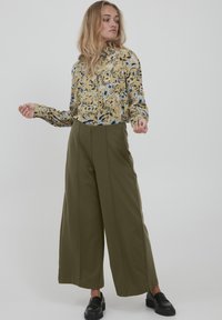 Chemise à boutons florale en jaune et bleu, assortie avec un pantalon large olive. Le modèle porte des mocassins noirs, mettant en avant une coupe décontractée et une texture.