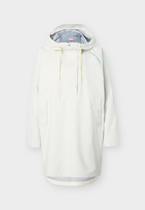 Impermeable blanco con capucha, cierre frontal con botones, cordones amarillos, bolsillos laterales y logo azul "Kari Traa" en el pecho.