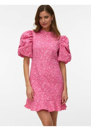 Kobieta ubrana w różową sukienkę w kwiaty z bufkami i falbaną u dołu, stojąca na jednolitym białym tle.