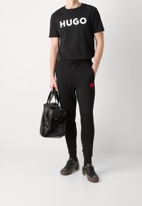 Hombre con camiseta negra HUGO, joggers negros con parche de logo rojo, zapatillas negras, sosteniendo una bolsa de duffel de cuero negro.