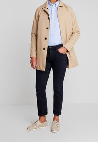 Beiger Trenchcoat mit braunen Knöpfen, hellblauem Hemd, dunklen Jeans und beigen Bootsschuhen. Lässiges Outfit mit einem klaren, maßgeschneiderten Look.