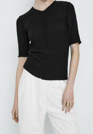 Personne portant un haut noir ajusté à manches courtes en tissu texturé et un pantalon blanc plissé taille haute, accessoirisé avec des bagues dorées.