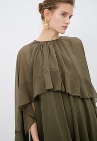 Olijfgroene chiffon jurk heeft een vloeiende, gelaagde ontwerp met een geraffineerde halslijn. Grote gouden statementoorbellen voegen contrast toe.