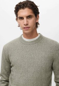 Joven con cabello corto y ondulado castaño, vistiendo un suéter de cuello redondo gris claro sobre una camisa blanca, posando frente a un fondo blanco liso.