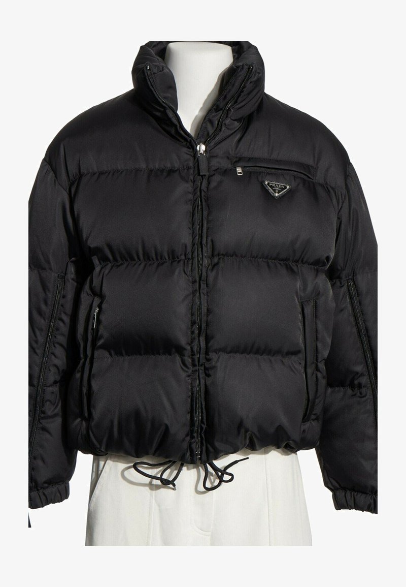 Doudoune noire Prada avec col montant, fermeture éclair devant, poche poitrine zippée et cordon de serrage à l'ourlet sur un buste de mannequin blanc.