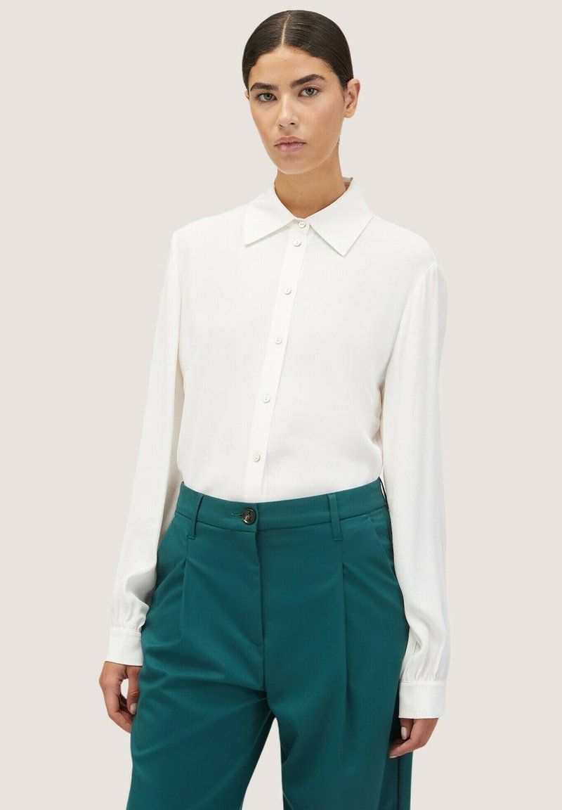 Blusa bianca a maniche lunghe con colletto a punta, chiusura a bottoni e tessuto testurizzato abbinata a pantaloni teal a vita alta con una piega frontale.