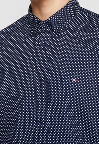 Camisa de manga corta azul marino con lunares blancos, cuello abotonado y pequeño logo bordado en el pecho. Tela de algodón suave.