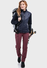 Dunkelblauer Pufferjacke mit schwarzen Ärmeln, bordeauxroten Slim-Fit-Hosen und schwarzen Wanderschuhen. Ausgestattet mit einem Rucksack und Trekkingstöcken.