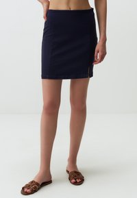 Gonna di jeans blu navy con design aderente, lunghezza mini e uno spacco laterale. Abbinata a sandali a fascia intrecciati marroni su uno sfondo chiaro.