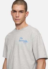 Camiseta de algodón gris con cuello redondo, mangas cortas y logo azul "Fila Sportslife Since 1974" impreso en el frente.