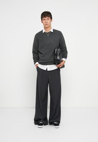 Grijze gebreide trui met een V-hals, wit overhemd eronder en brede antracietkleurige broek. Houdt een zwarte gestructureerde tas vast en draagt zwarte sneakers.